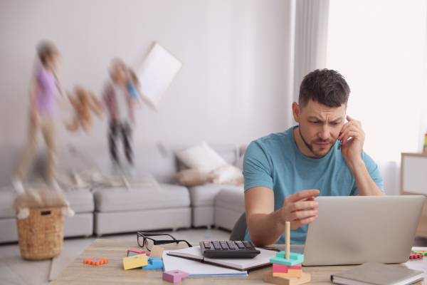 Vater am Schreibtisch verzweifelt, Kinder spielen im Hintergrund